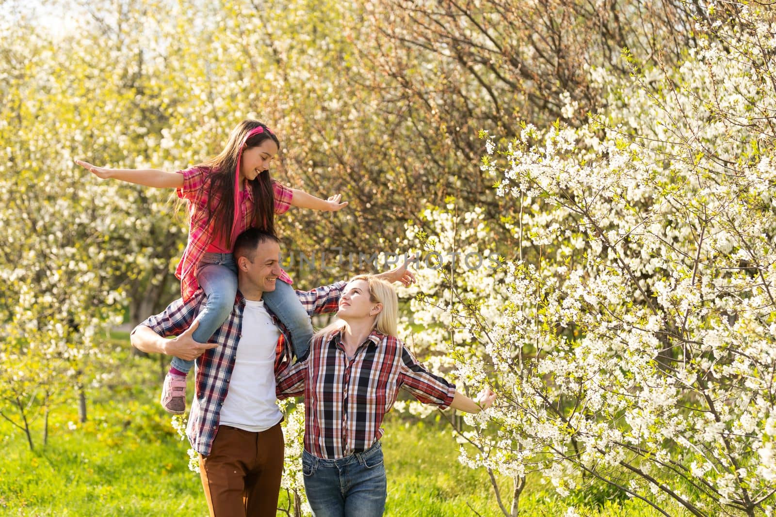 Outdoor portrait of happy young family playing in spring park under blooming tree, lovely couple with little child having fun in sunny garden.
