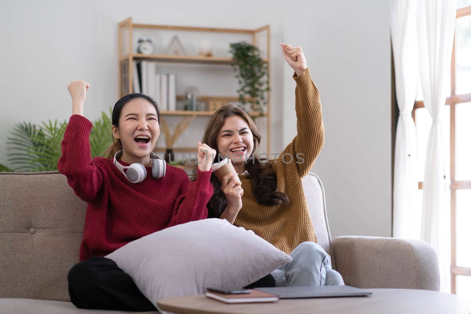 Two Young womanWatching TV Shaking Fists In Joy Celebrating Victory Of Favorite Sport Team Sitting On Couch In Living Room At Home. Weekend Leisure, Television Show And Entertainment Concept.
