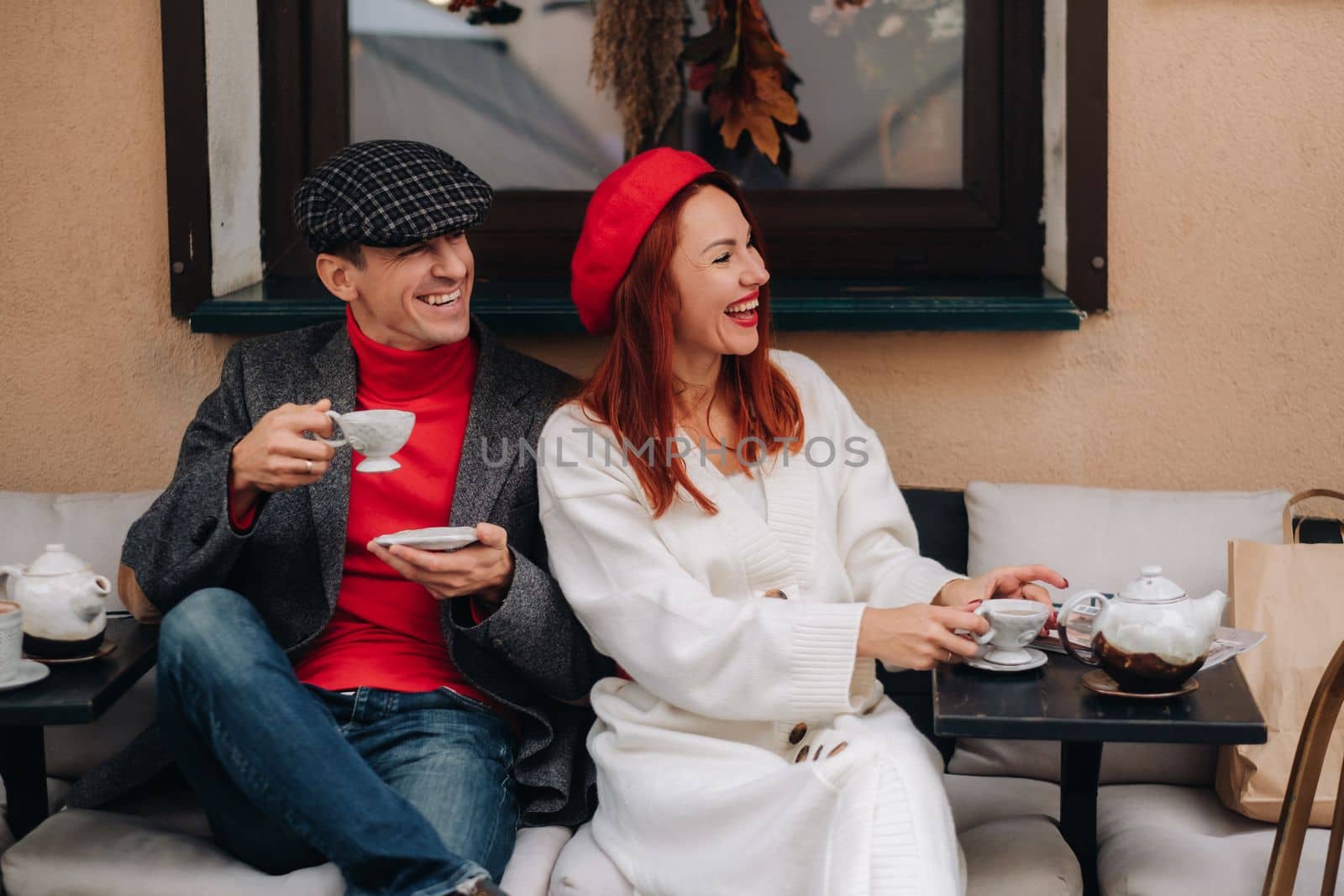 A happy stylish couple drinks coffee and smiles while sitting in a cafe on the street.