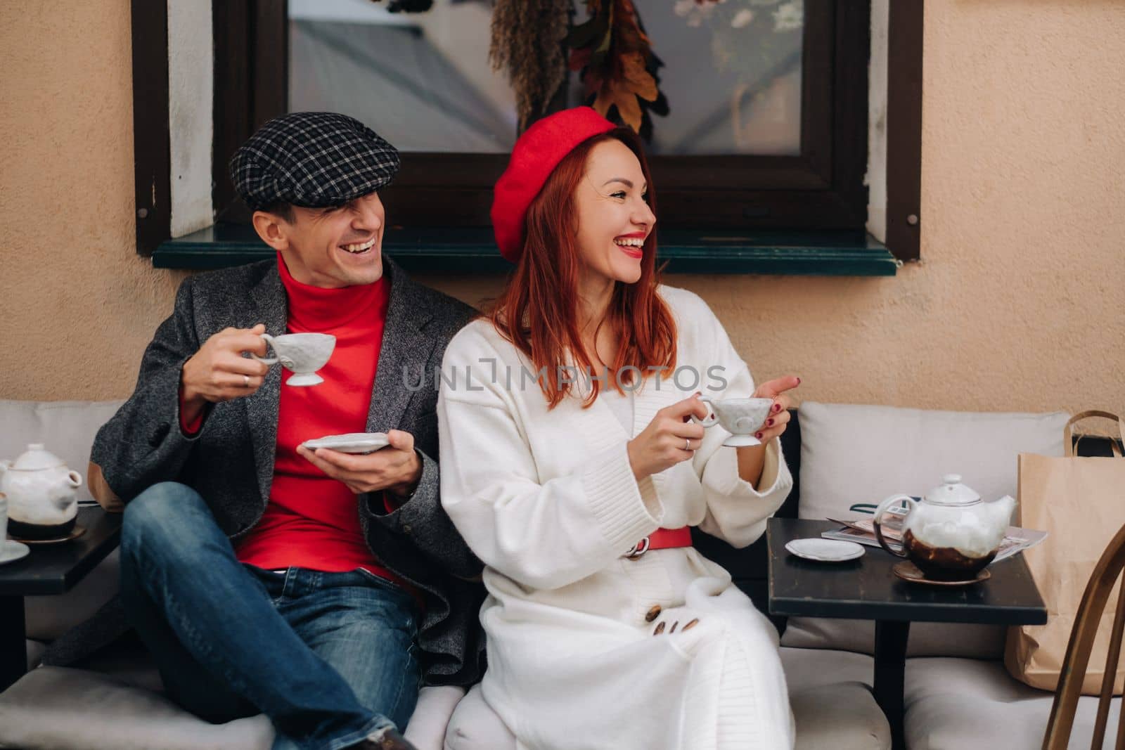 A happy stylish couple drinks coffee and smiles while sitting in a cafe on the street by Lobachad