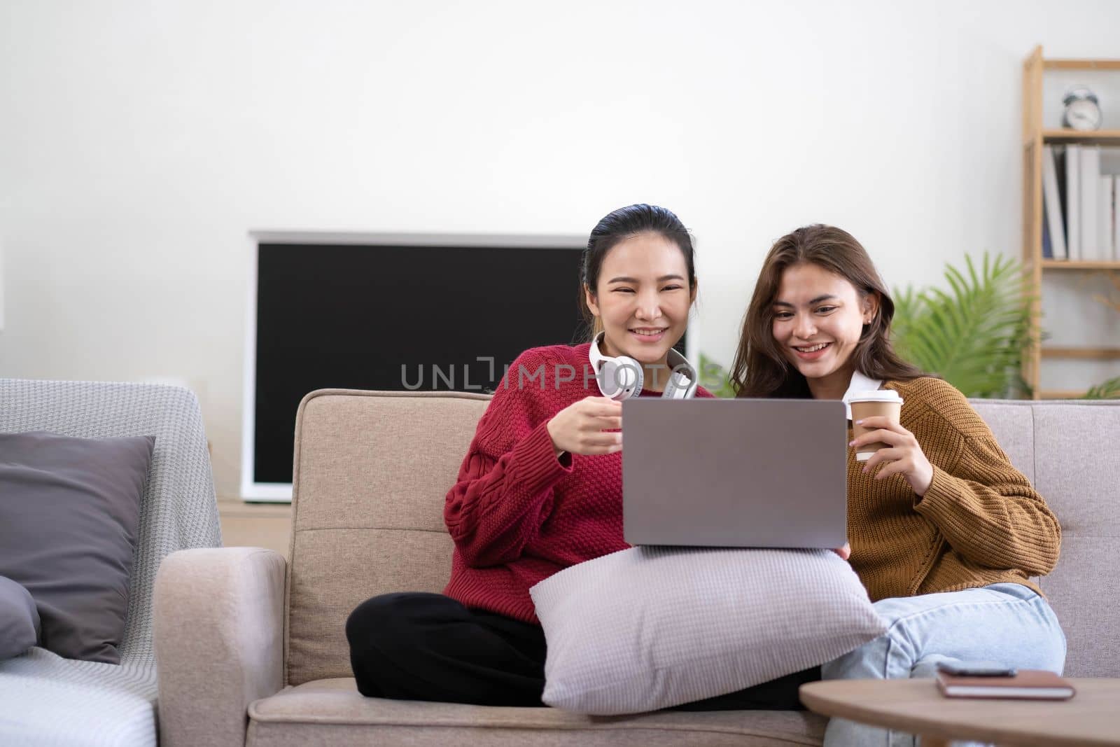 Two asian young woman happy smiling and using computer laptop on couch in living room at home by wichayada