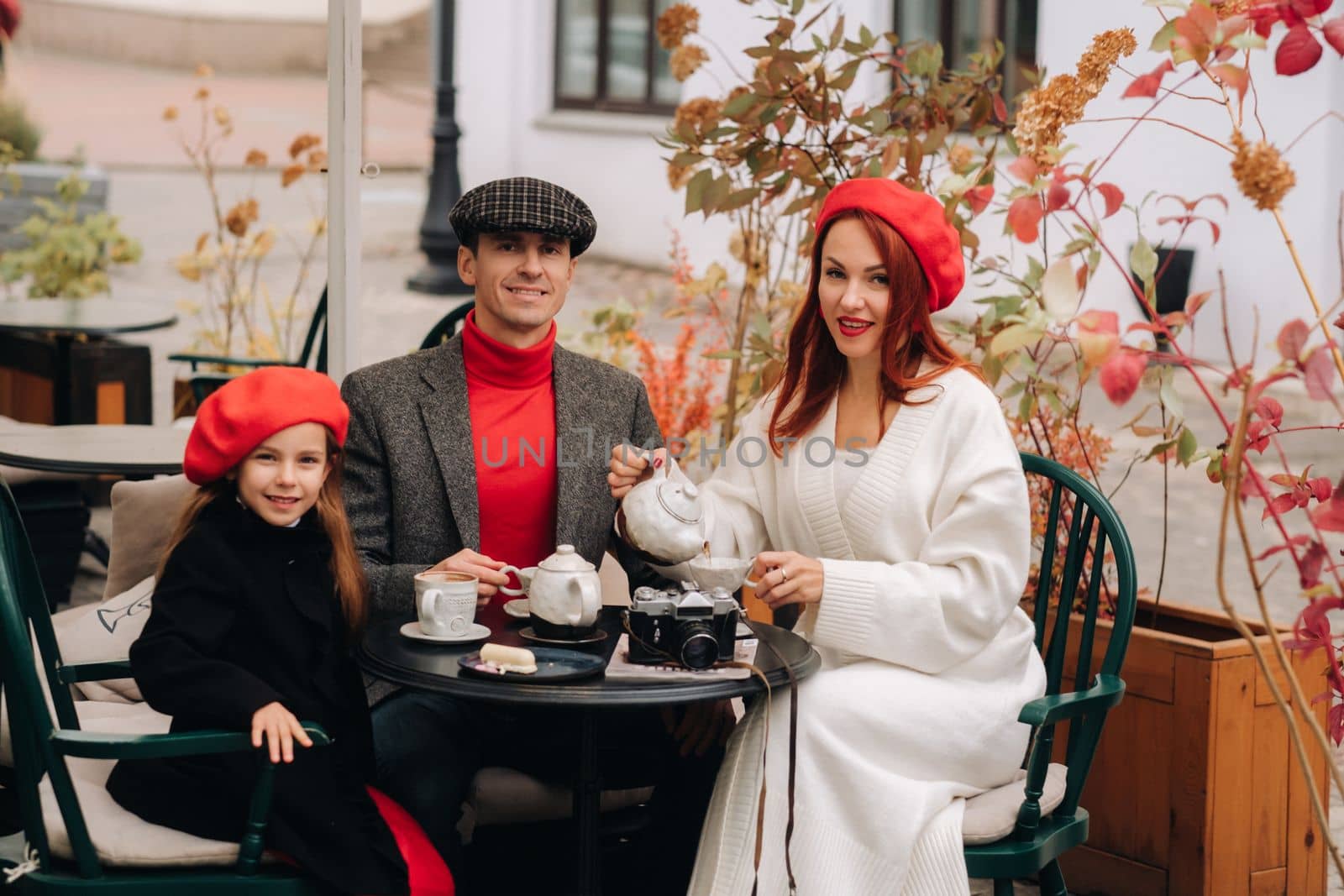 A stylish family gathered together in a cafe on the street. Mom, dad, little daughter drink tea, eat cakes. They are happy together. The concept of a happy family dinner.