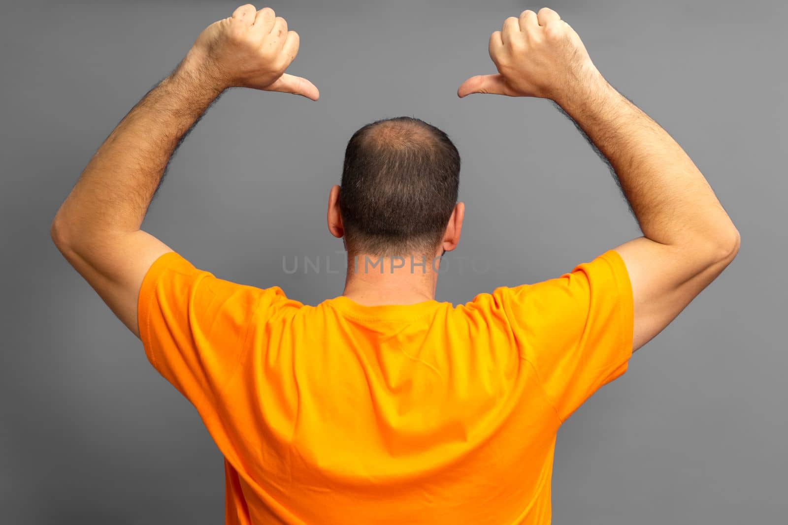Proud man in orange t-shirt with his back turned pointing with his thumbs at his receding bald spot, isolated on gray background
