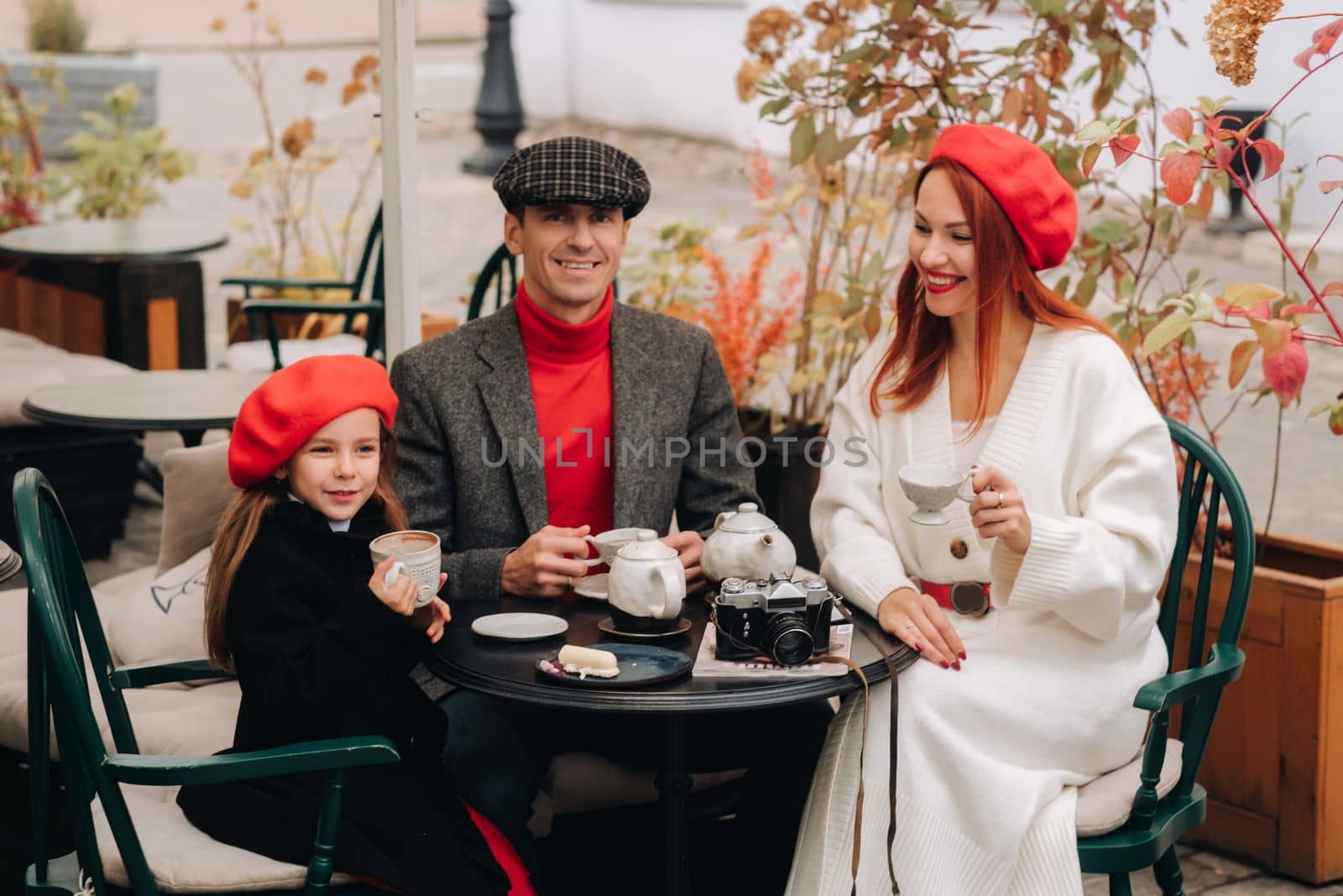 A stylish family gathered together in a cafe on the street. Mom, dad, little daughter drink tea, eat cakes. They are happy together. The concept of a happy family dinner.