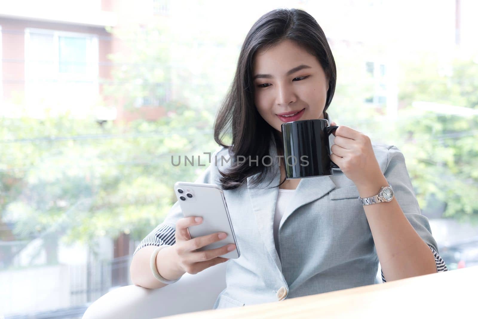 Beautiful young Asian businesswoman smiling holding a coffee mug and smartphone working at the office..