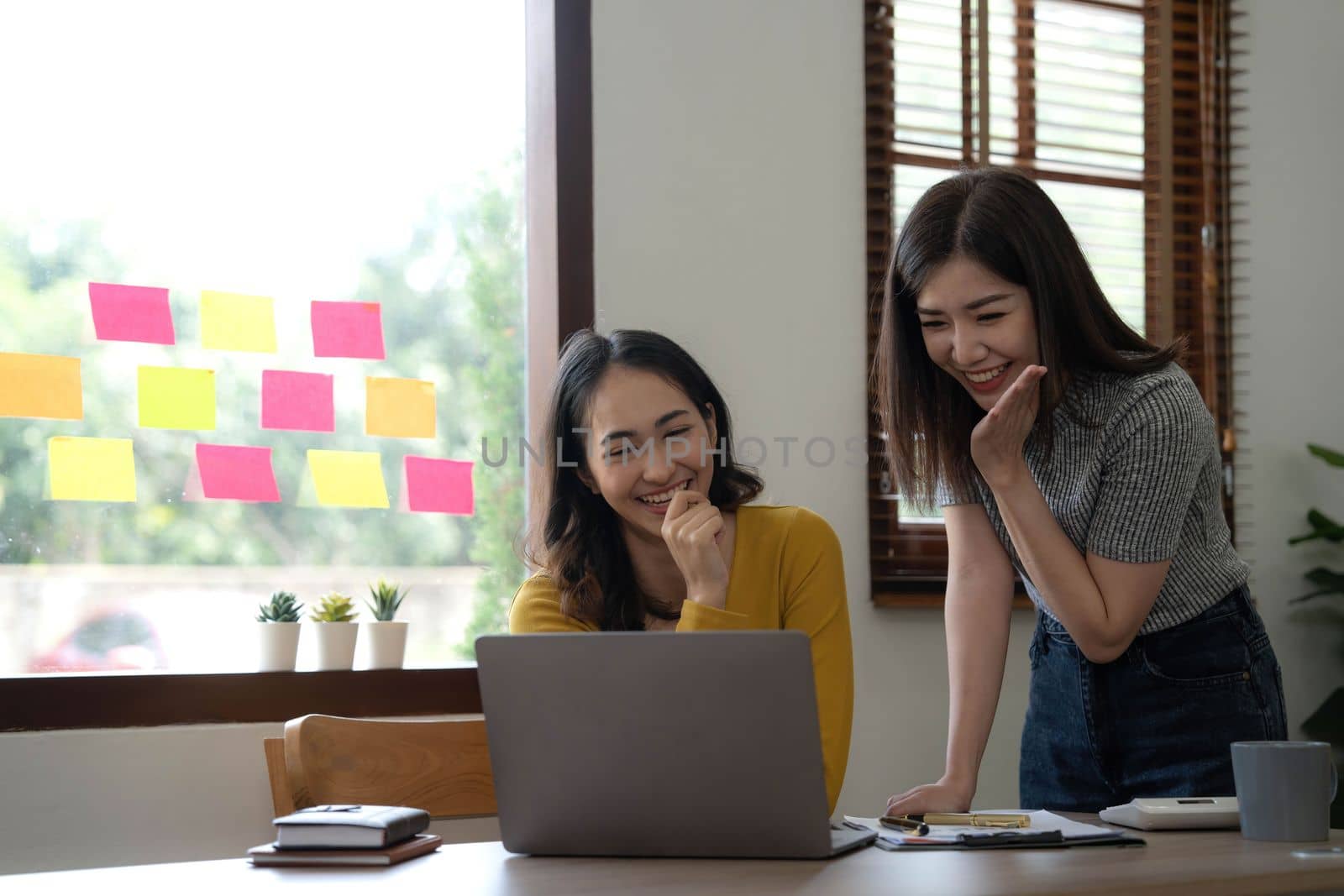 Two young Asian businesswomen show joyful expression of success at work smiling happily with a laptop computer in a modern office. by wichayada