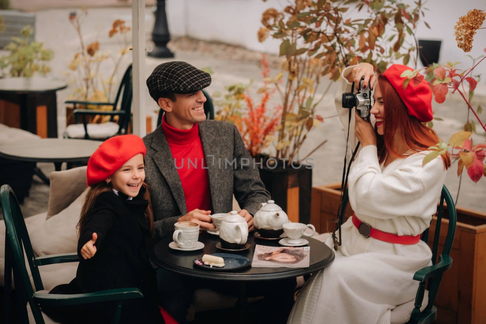 A stylish family of three is sitting at a table outside in a cafe and drinking coffee. Dad, mom and daughter in the autumn city.