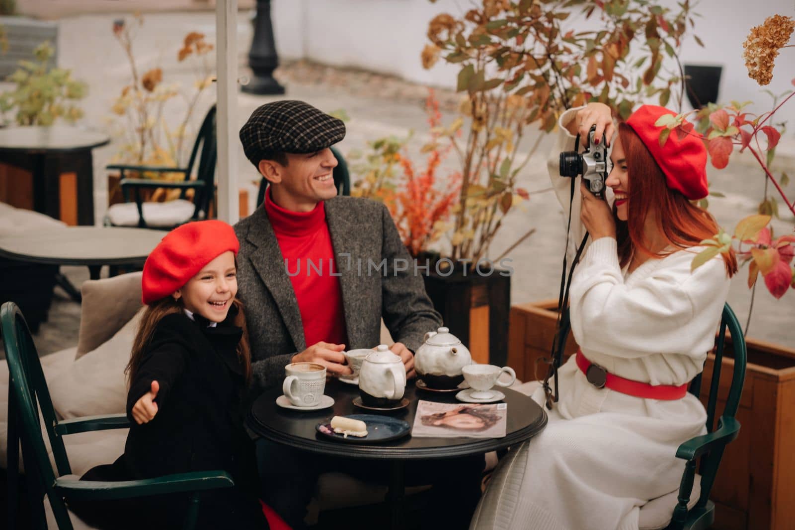 A stylish family of three is sitting at a table outside in a cafe and drinking coffee. Dad, mom and daughter in the autumn city by Lobachad