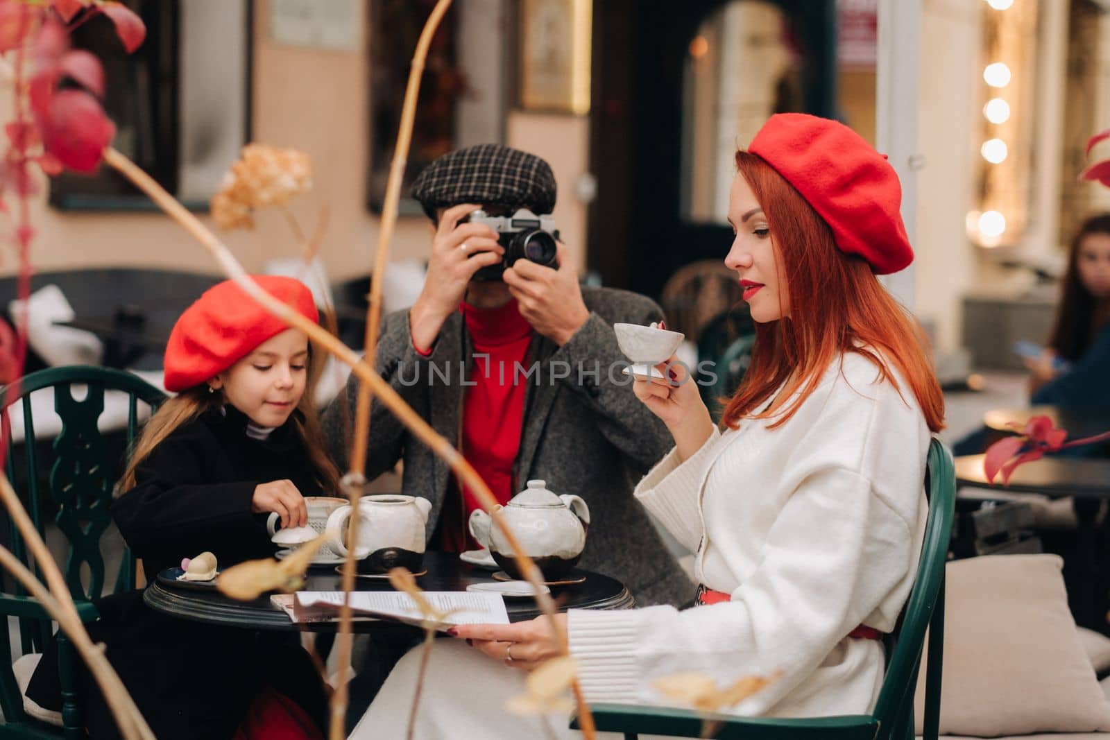 A stylish family of three is sitting at a table outside in a cafe and drinking coffee. Dad takes pictures of mom in the autumn city.