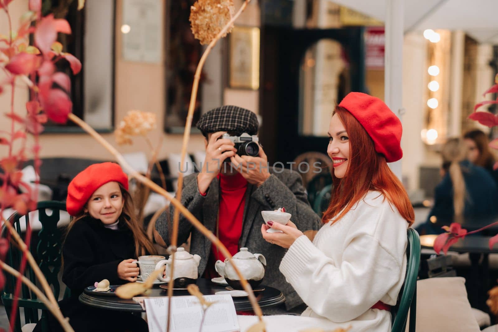A stylish family of three is sitting at a table outside in a cafe and drinking coffee. Dad takes pictures of mom in the autumn city.