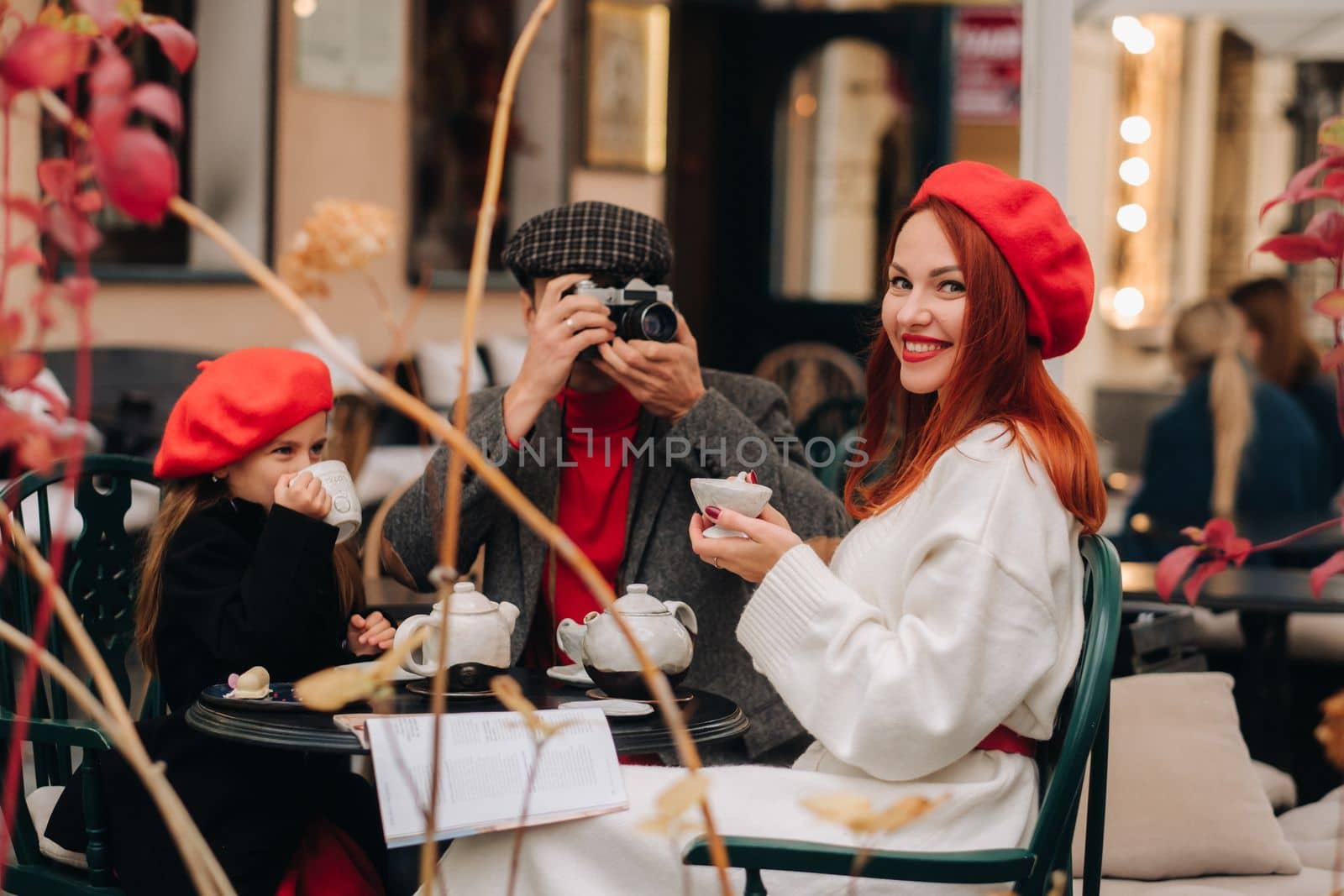 A stylish family of three is sitting at a table outside in a cafe and drinking coffee. Dad takes pictures of mom in the autumn city.