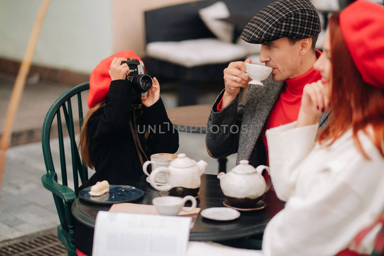 A stylish family of three is sitting at a table outside in a cafe and drinking coffee. Dad, mom and daughter in the autumn city by Lobachad