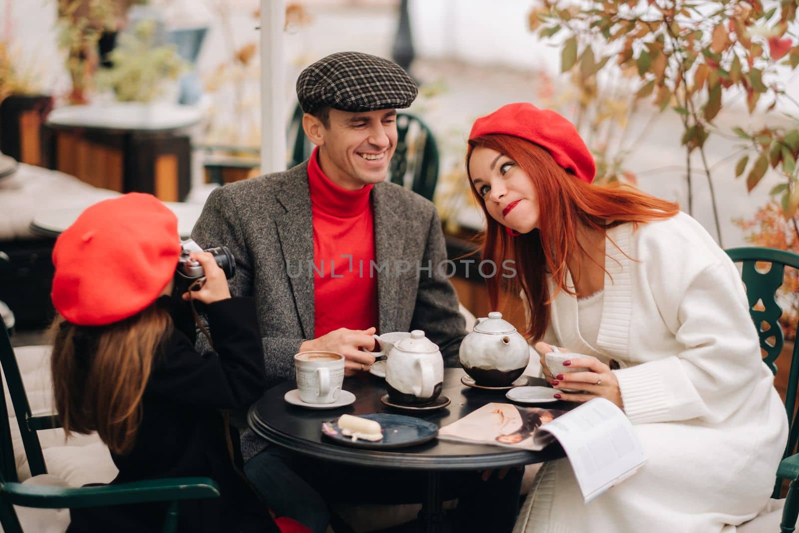 A stylish family of three is sitting at a table outside in a cafe and drinking coffee. Dad, mom and daughter in the autumn city.