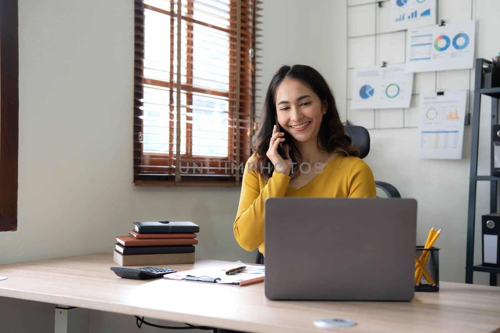 Smiling beautiful Asian businesswoman analyzing chart and graph showing changes on the market and holding smartphone at office..