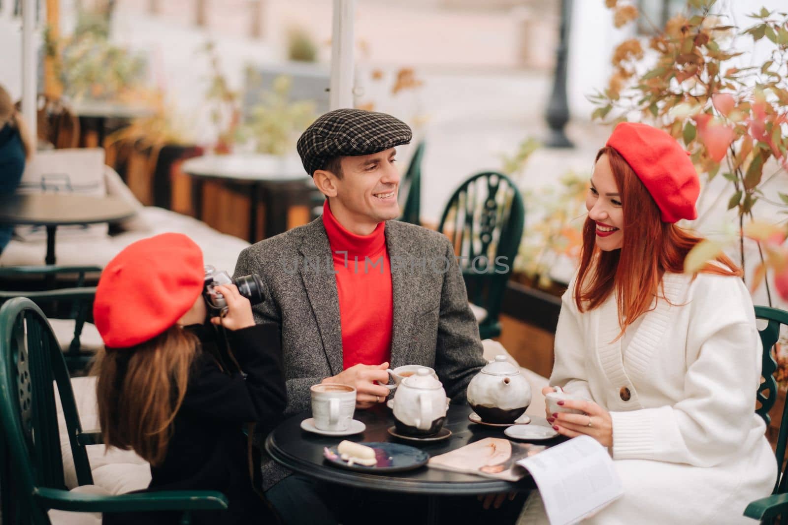 A stylish family of three is sitting at a table outside in a cafe and drinking coffee. Dad, mom and daughter in the autumn city by Lobachad