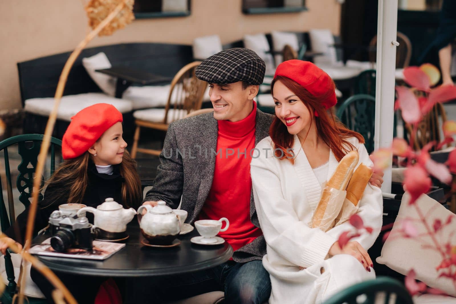 A stylish family gathered together in a cafe on the street. Mom, dad, little daughter drink tea, eat cakes. They are happy together. The concept of a happy family dinner by Lobachad