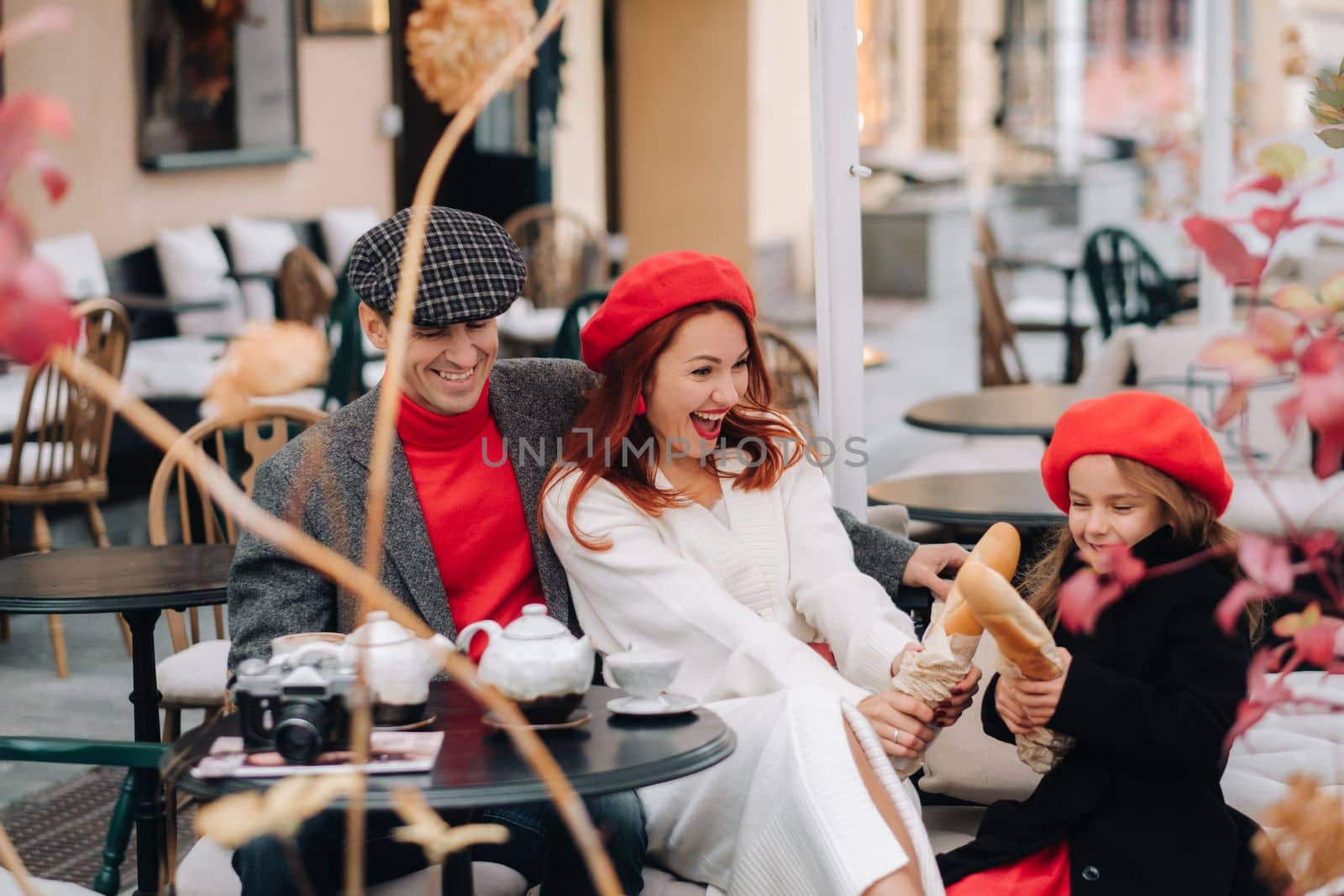 A stylish family of three is sitting at a table outside in a cafe and drinking coffee. Dad, mom and daughter in the autumn city.