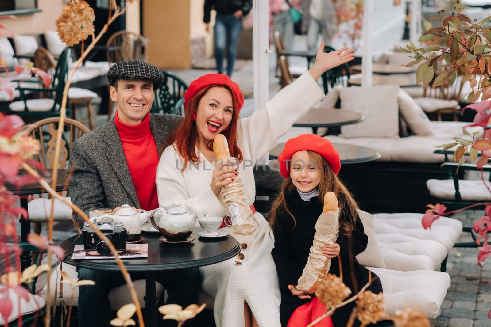 A stylish family of three is sitting at a table outside in a cafe and drinking coffee. Dad, mom and daughter in the autumn city.