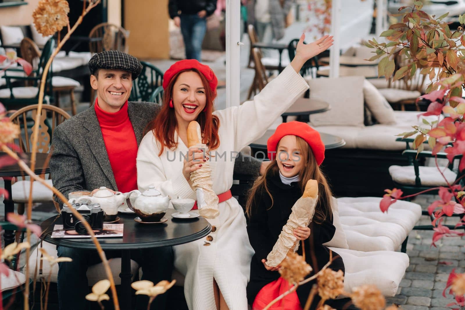 A stylish family of three is sitting at a table outside in a cafe and drinking coffee. Dad, mom and daughter in the autumn city.