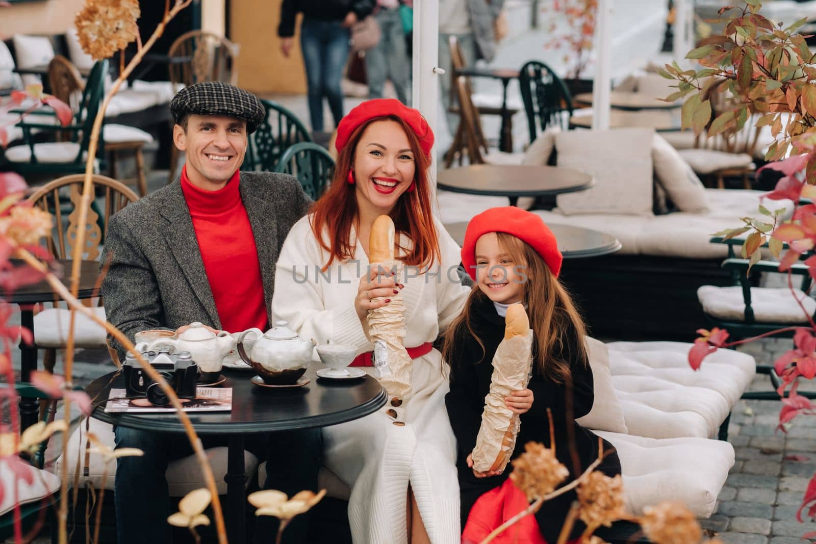 A stylish family of three is sitting at a table outside in a cafe and drinking coffee. Dad, mom and daughter in the autumn city by Lobachad