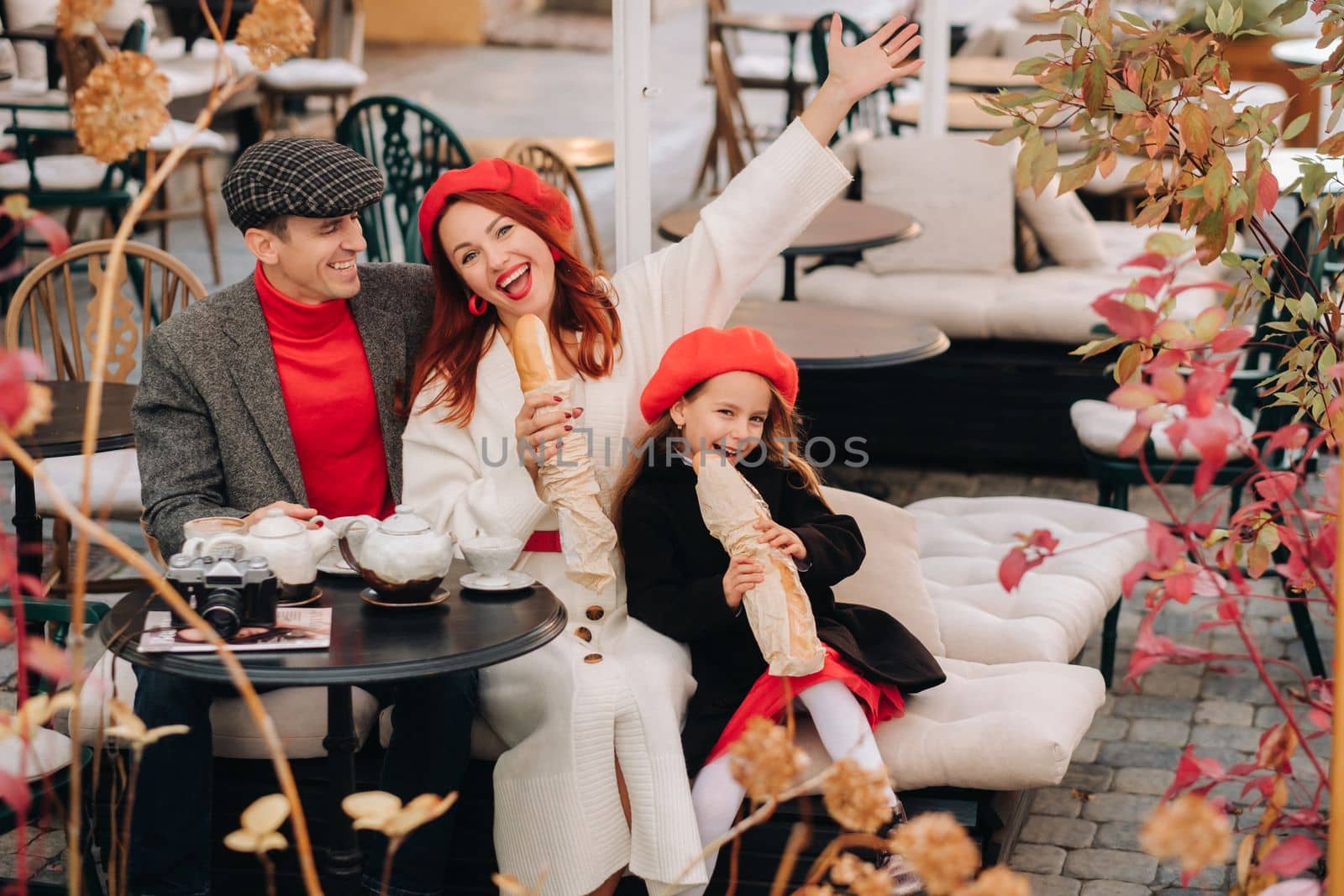 A stylish family of three is sitting at a table outside in a cafe and drinking coffee. Dad, mom and daughter in the autumn city.