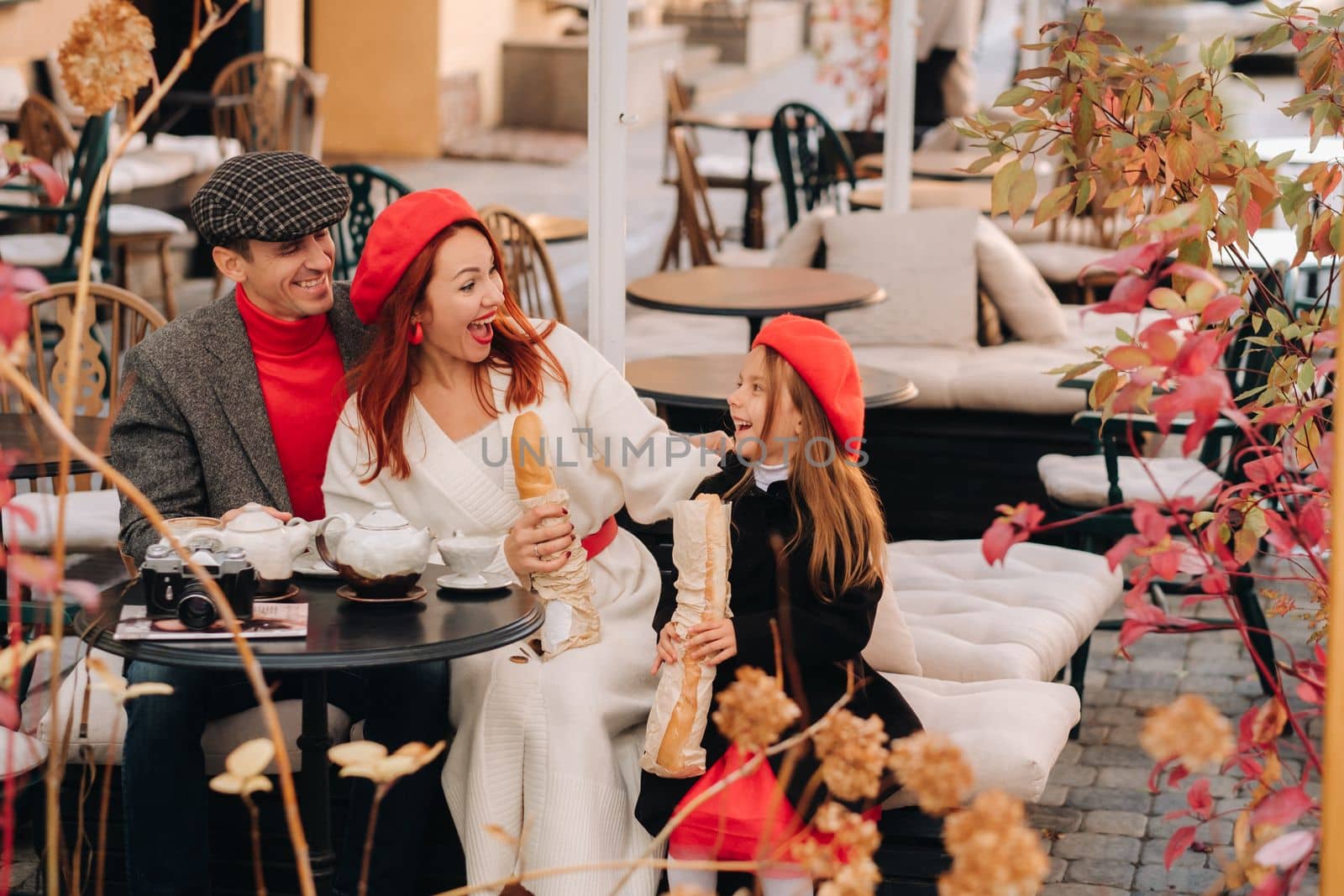 A stylish family of three is sitting at a table outside in a cafe and drinking coffee. Dad, mom and daughter in the autumn city.