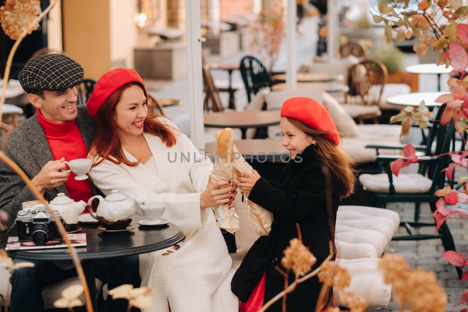 A stylish family of three is sitting at a table outside in a cafe and drinking coffee. Dad, mom and daughter in the autumn city.