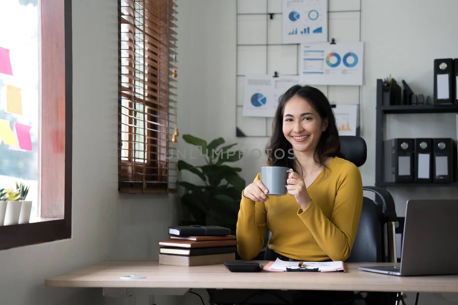 Beautiful young Asian businesswoman smiling holding a coffee mug and laptop working at the office..