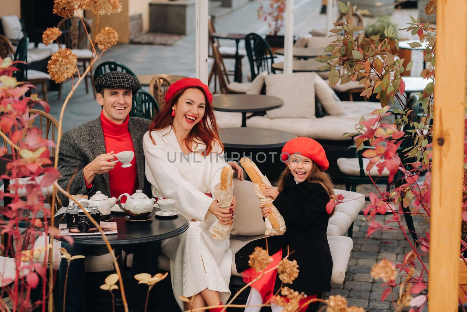 A stylish family of three is sitting at a table outside in a cafe and drinking coffee. Dad, mom and daughter in the autumn city.