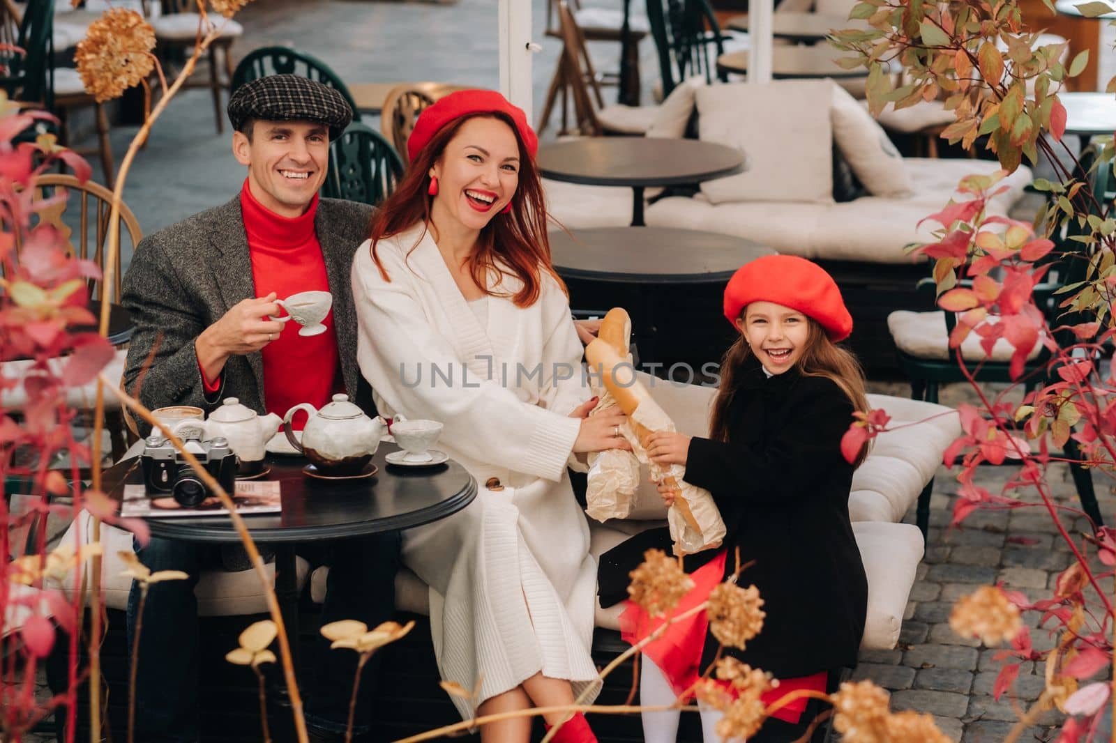 A stylish family of three is sitting at a table outside in a cafe and drinking coffee. Dad, mom and daughter in the autumn city.