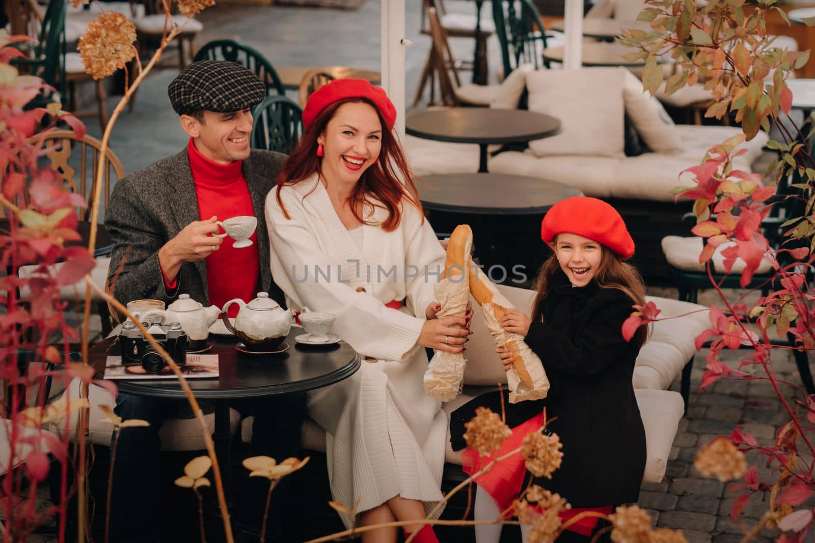 A stylish family of three is sitting at a table outside in a cafe and drinking coffee. Dad, mom and daughter in the autumn city.