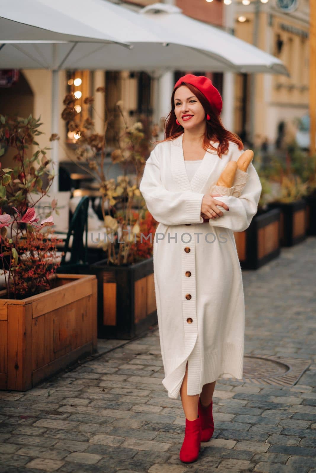 Portrait of a pretty woman in a red beret and a white cardigan with baguettes in her hands strolling through the autumn city by Lobachad