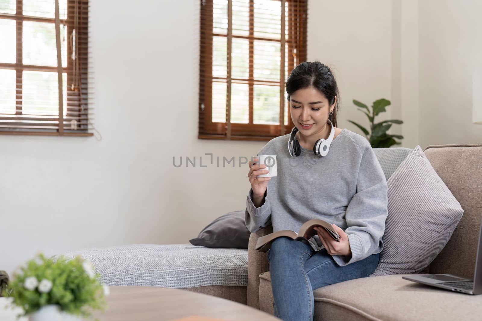 Young beautiful asian woman student sitting on sofa reading book at home while using laptop for online learning