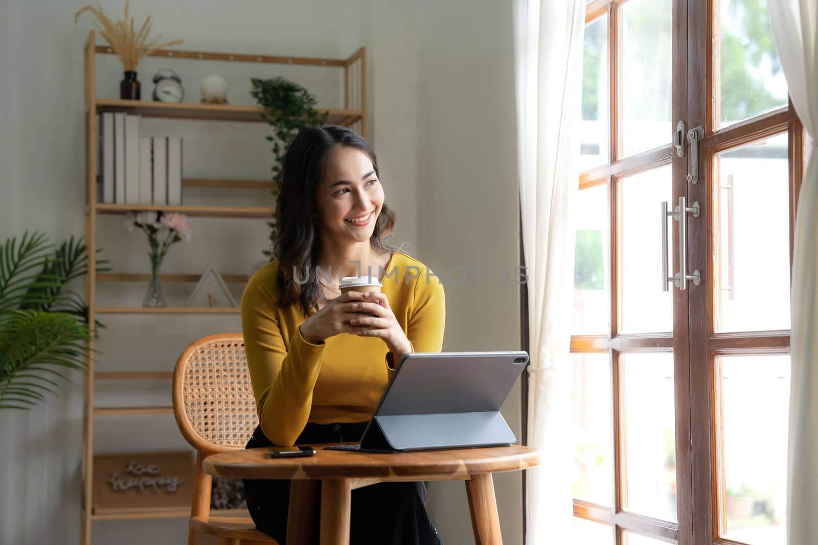 Beautiful Asian woman is using laptop, drinking coffee and smiling while sitting by the window at home...