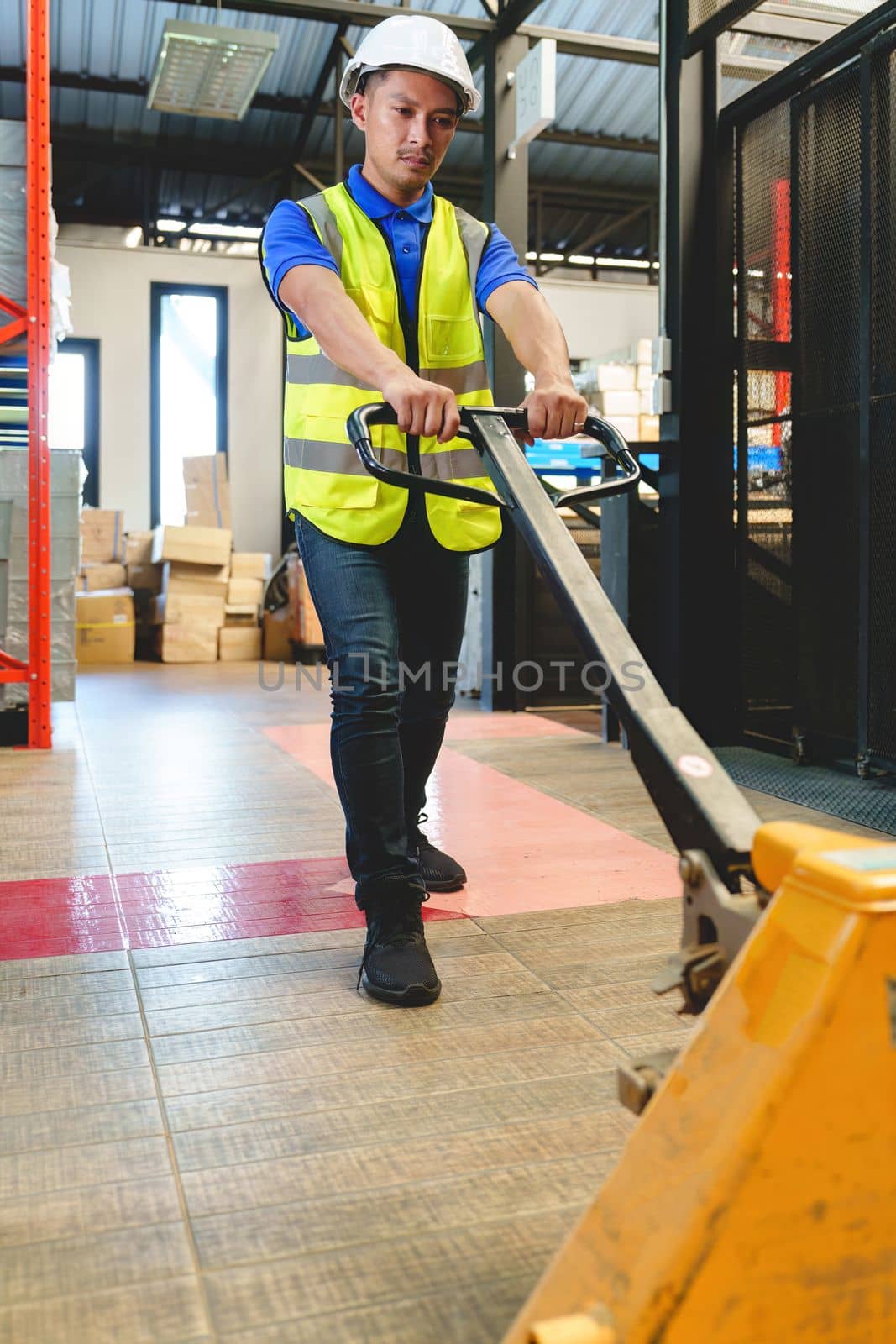 Asian male warehouse worker pulling a pallet truck. Worker working with hand pallet truck unloading cargo boxes on pallet at the warehouse. by nateemee