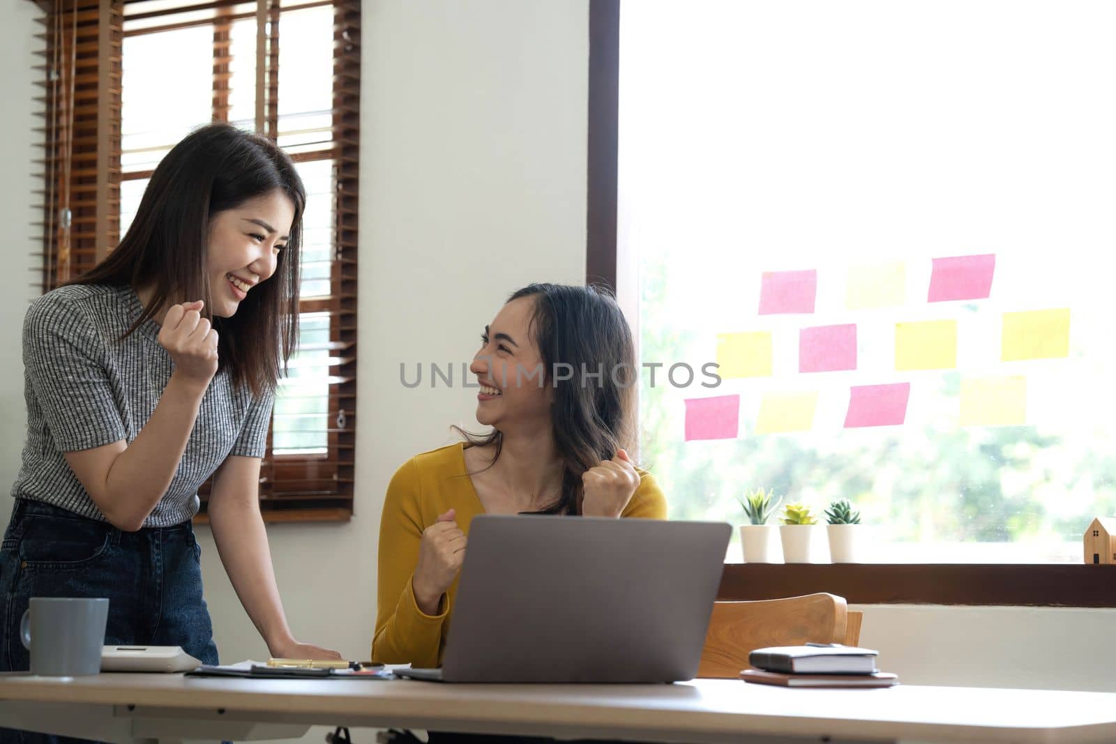 Two young Asian businesswomen show joyful expression of success at work smiling happily with a laptop computer in a modern office. by wichayada