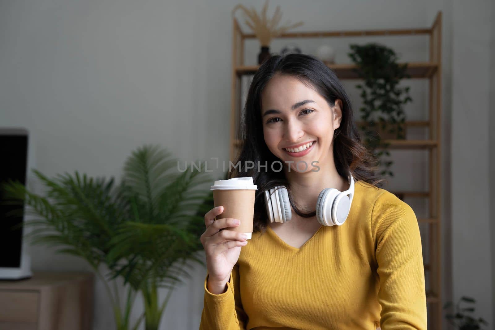 Beautiful Asian woman is using laptop, drinking coffee and smiling while sitting by the window at home...