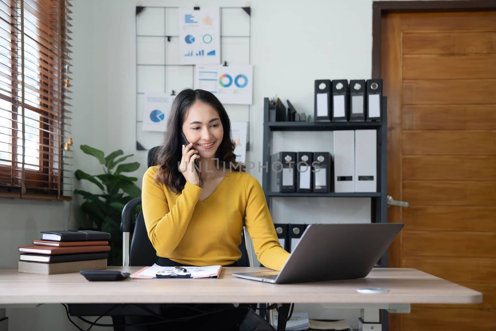 Smiling beautiful Asian businesswoman analyzing chart and graph showing changes on the market and holding smartphone at office..