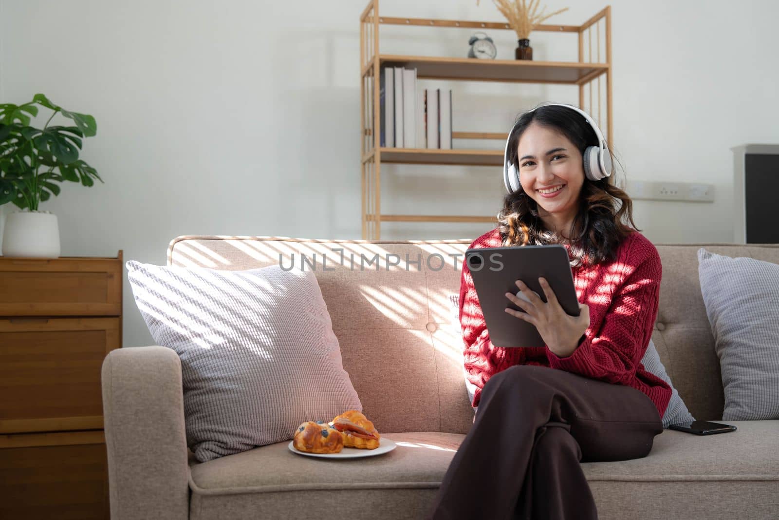 Beautiful woman asian wearing sweater listening music on line with a tablet sitting on a sofa in the living room at home