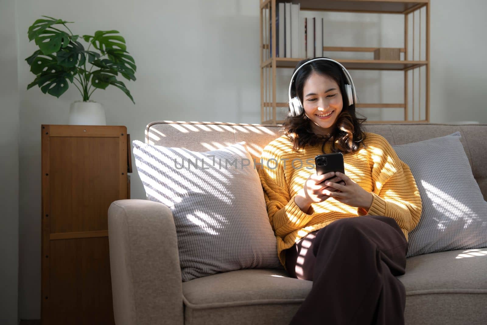 Young Asian woman listening music from headphones and writing note for her work idea in diary book.She sitting on grey sofa in living room..