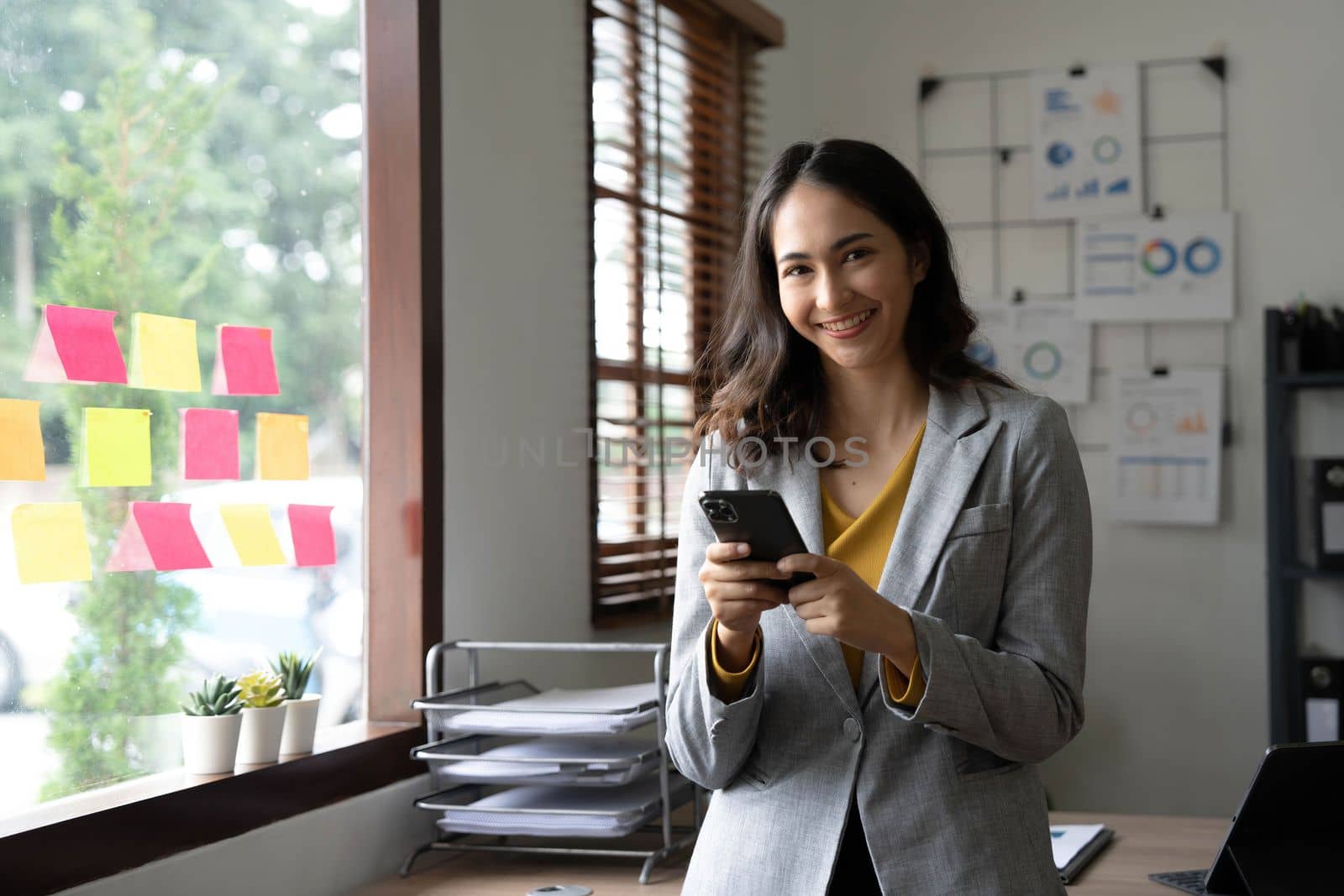 Smiling beautiful Asian businesswoman analyzing chart and graph showing changes on the market and holding smartphone at office..