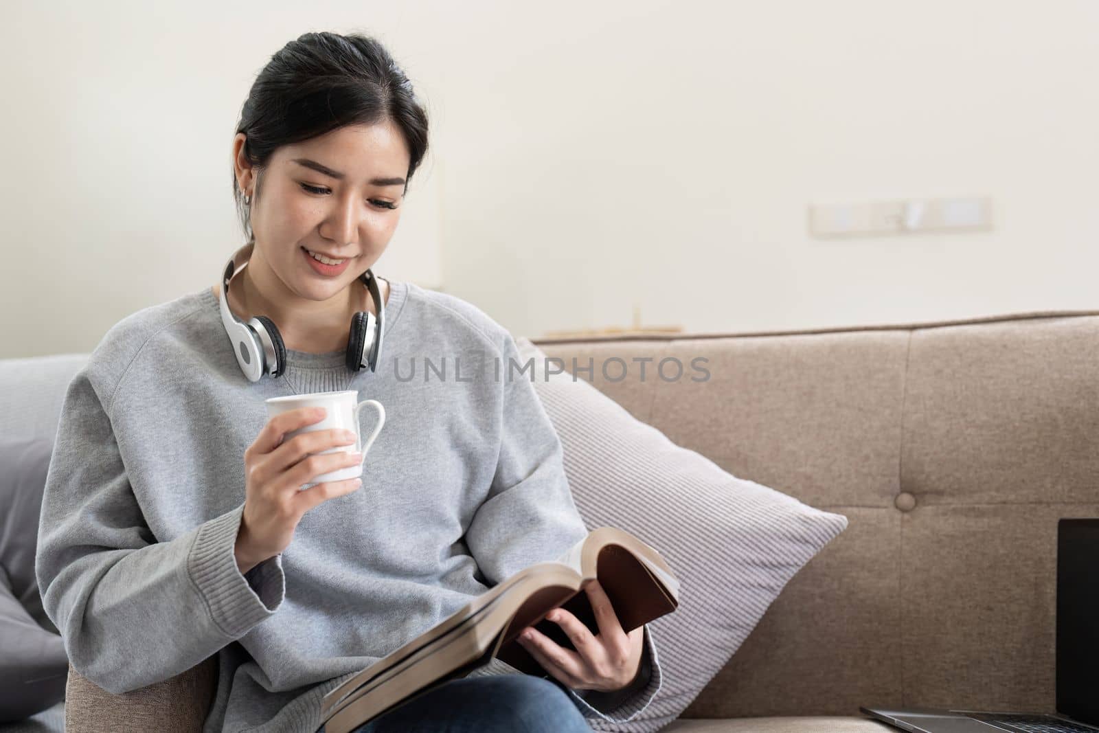 Young beautiful asian woman student sitting on sofa reading book at home while using laptop for online learning.