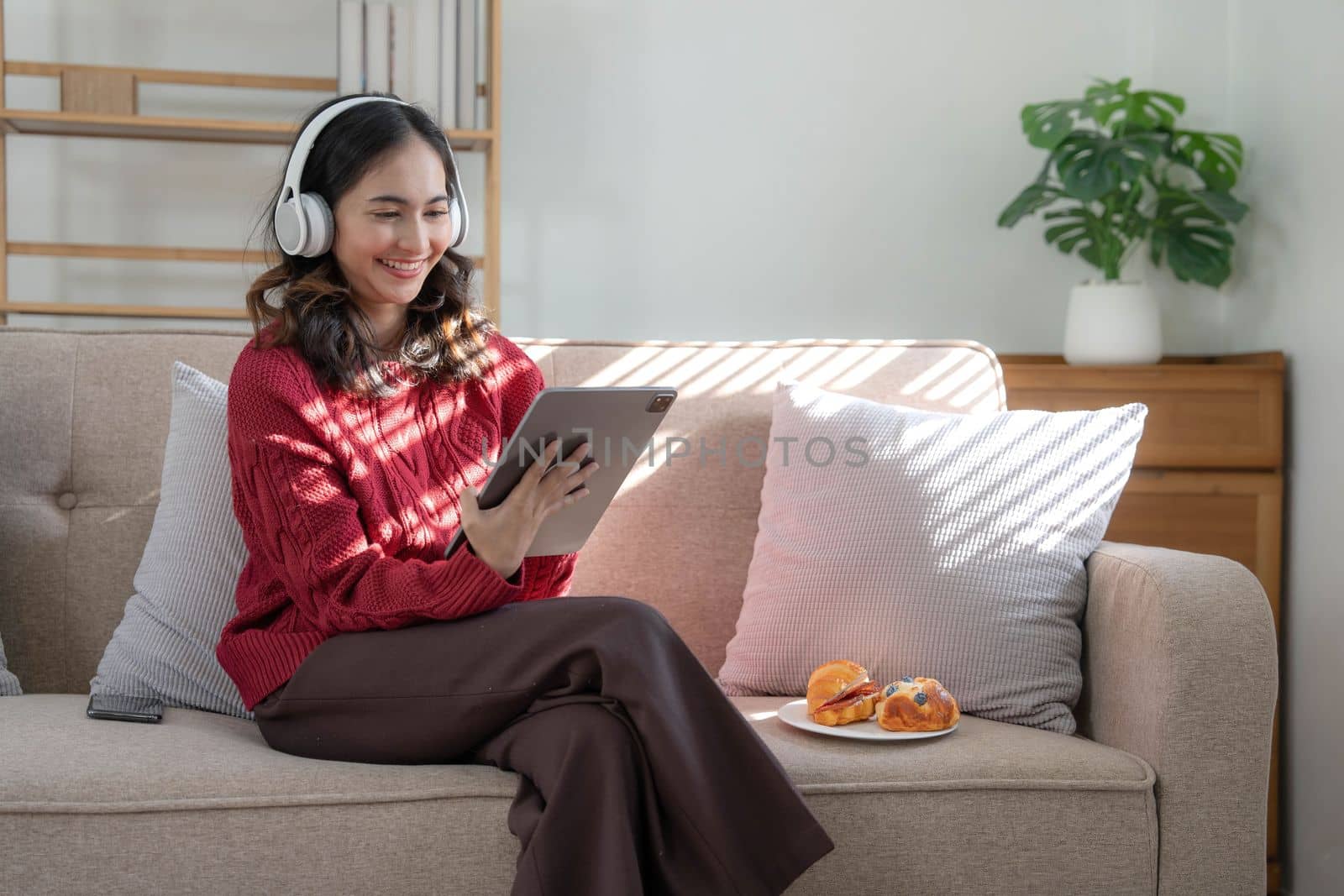 Beautiful woman asian wearing sweater listening music on line with a tablet sitting on a sofa in the living room at home