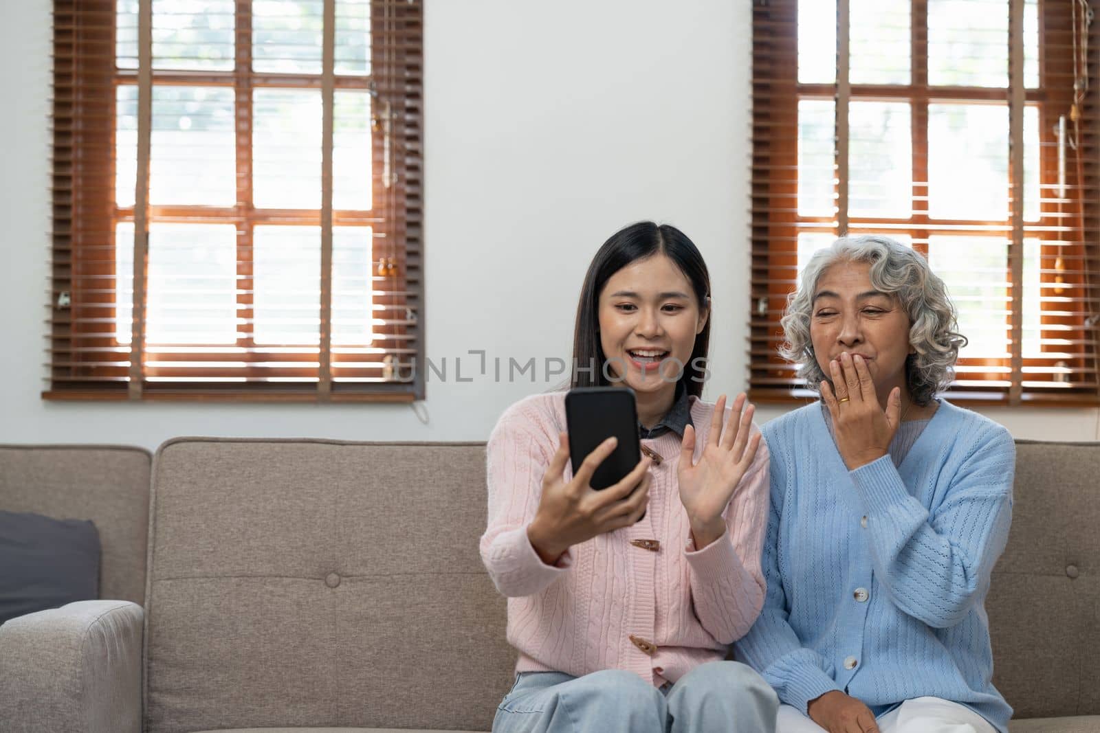 Grandmother and granddaughter sitting on sofa and having fun. Happy elderly woman learning to make video call on mobile phone by nateemee