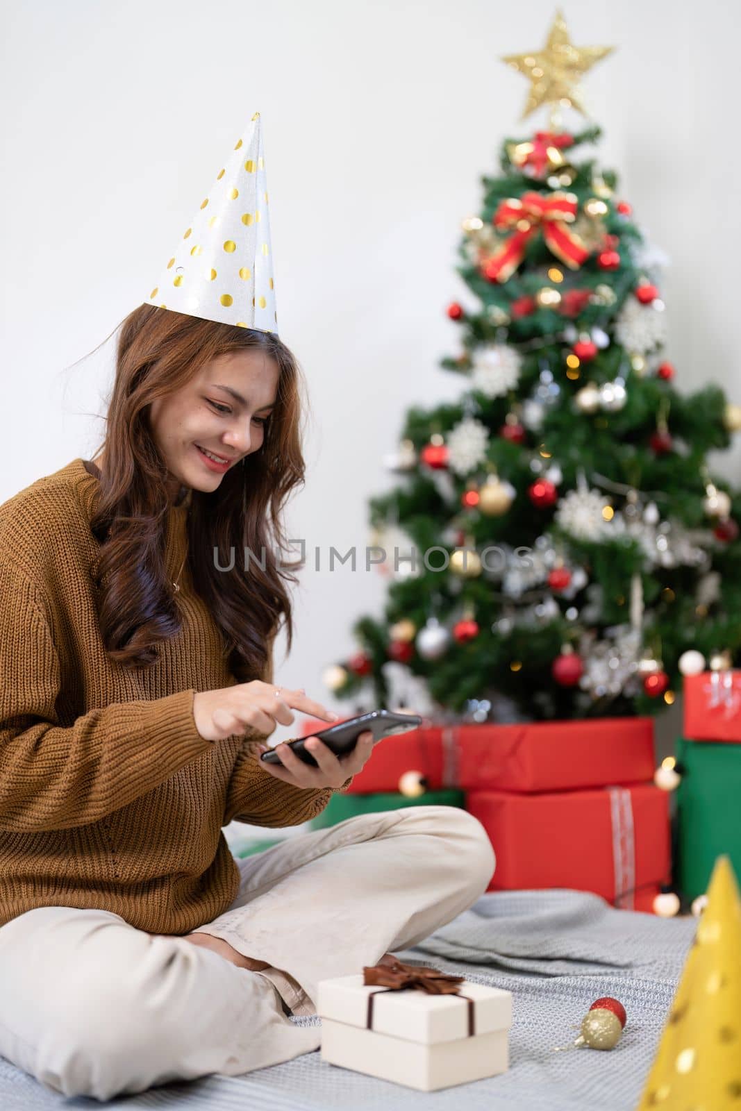 Portrait woman reading typing holiday congrats message use smartphone at festive decorated Christmas tree. Smiling beautiful female chatting on mobile phone at Xmas spruce with positive emotion.