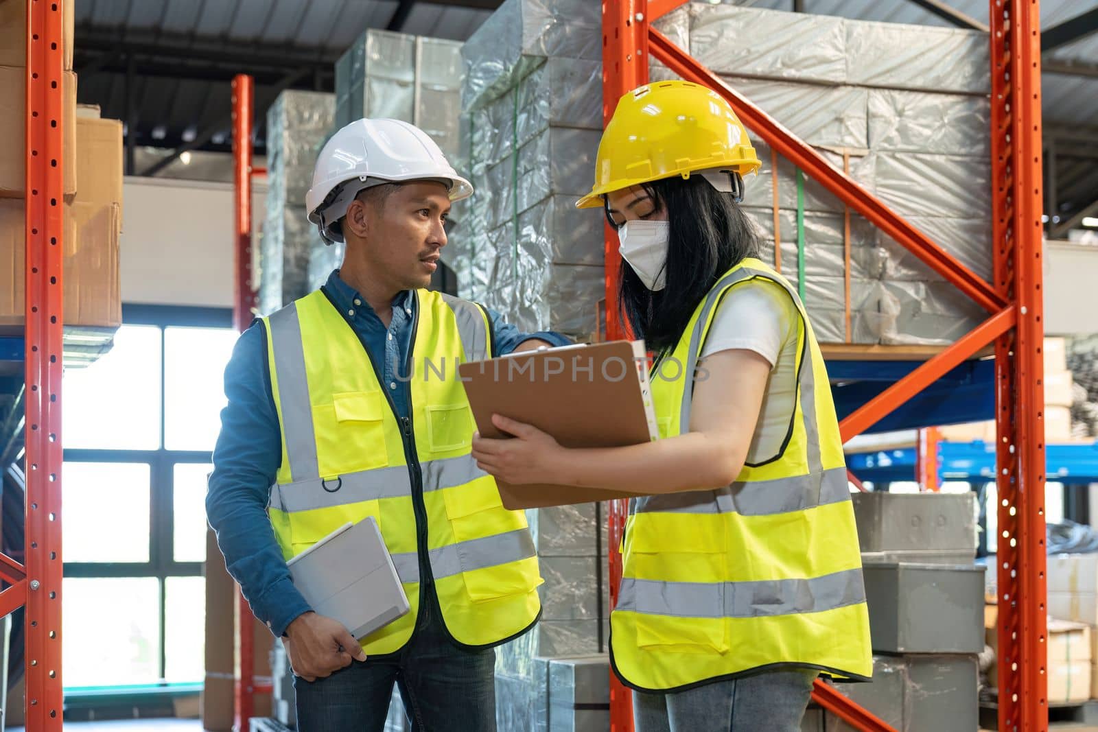 Two smile warehouse workers in uniforms and yellow helmets on heads standing and talking about job. adviser.