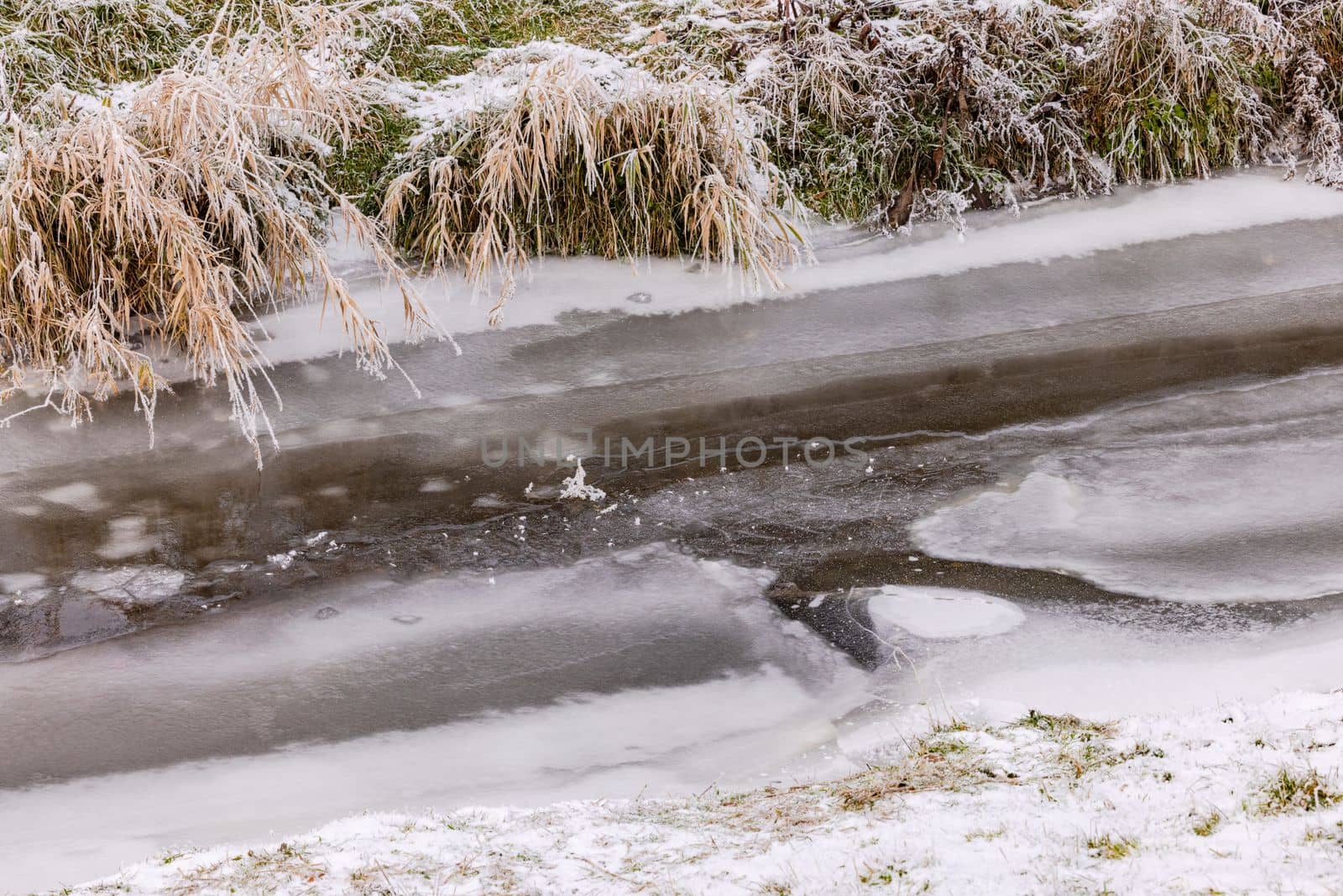 A completely frozen stream with ice and snow in winter in a rural landscape with grass on the bank, Germany