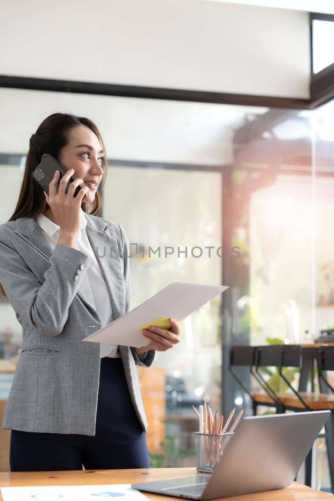 Happy young asian business woman wearing suit holding mobile phone standing in her workstation office by wichayada