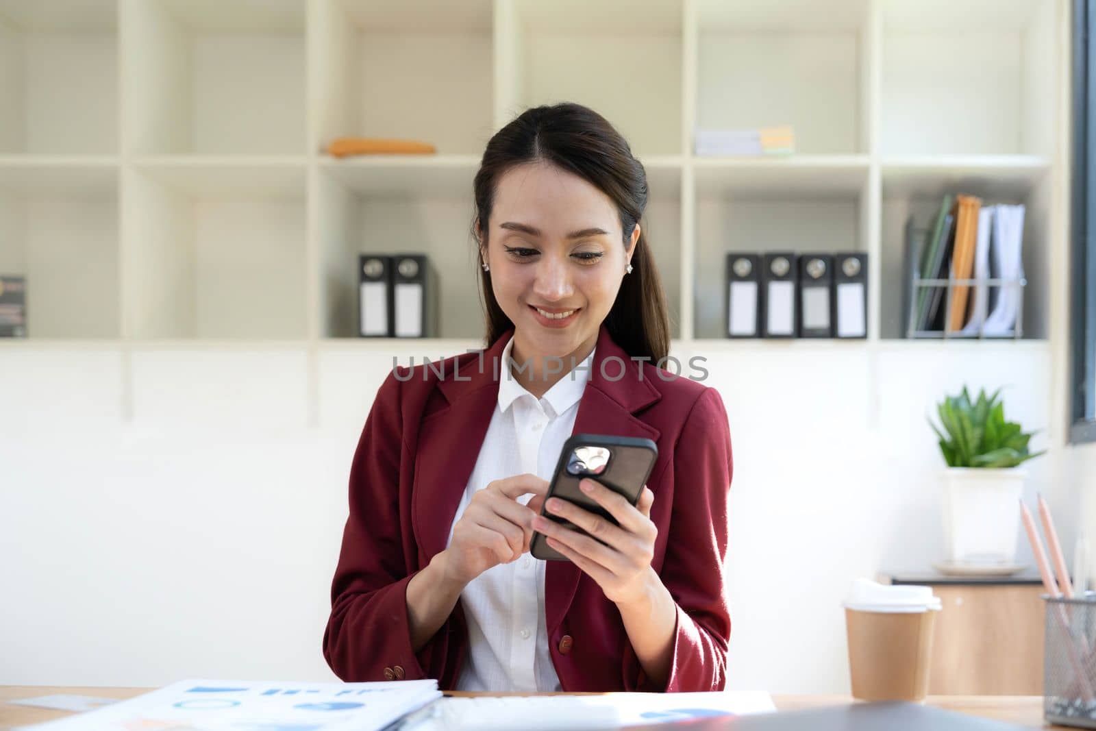Smiling beautiful Asian businesswoman analyzing chart and graph showing changes on the market and holding smartphone at office..
