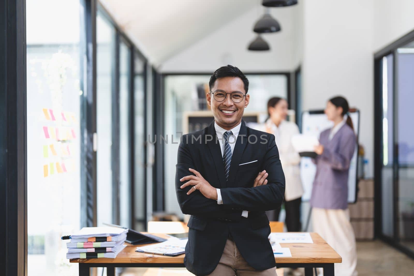 Portrait of success asian male office worker in formal business suits smiling at camera in office with blurry colleagues working in office as background